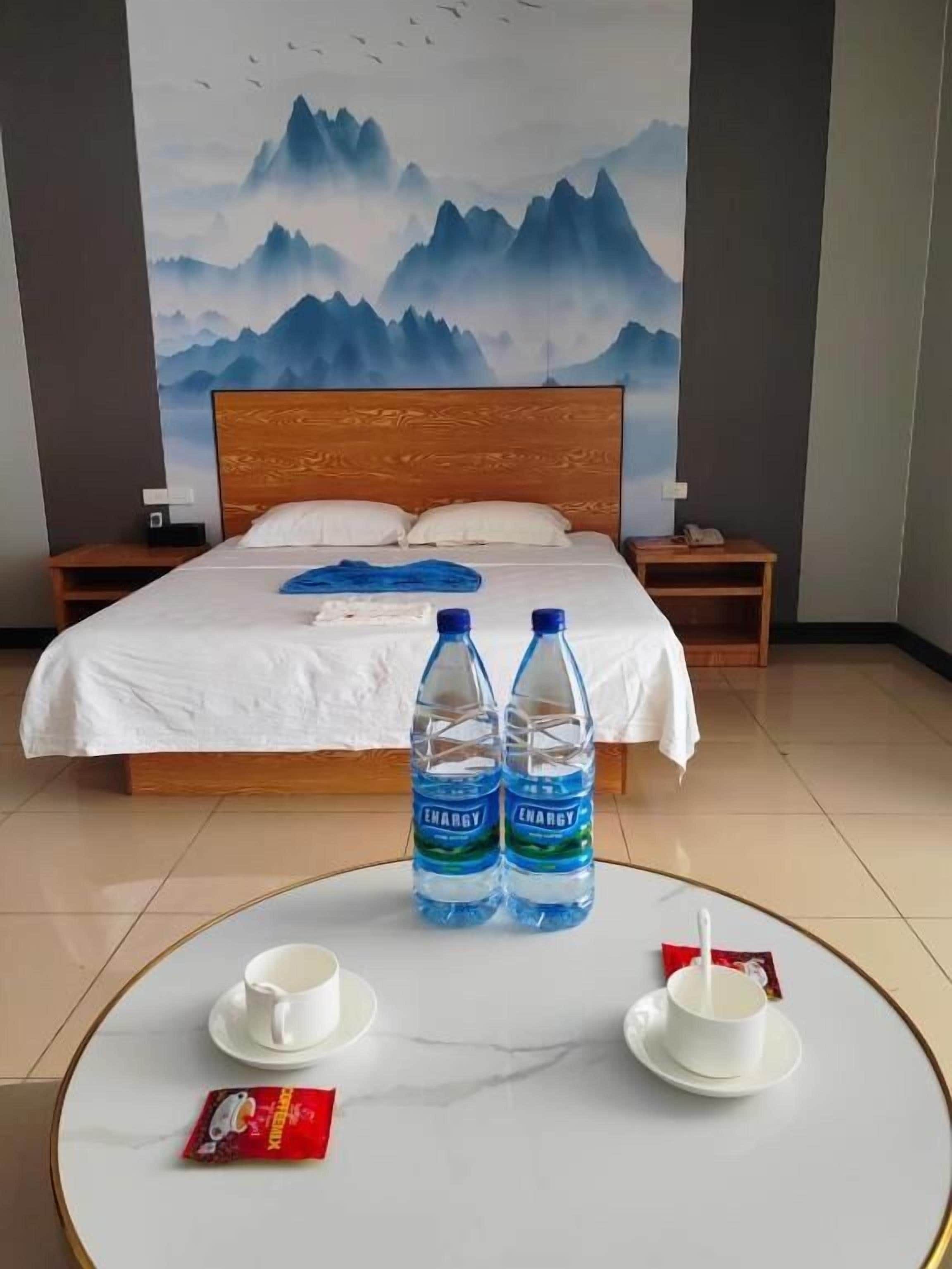 A hotel room with a bed, two bedside tables, a round coffee table with cups and a packet, and a large wall painting of mountains.