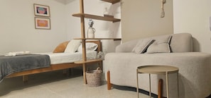 A bedroom with a bed, a sofa, a wooden shelf unit, and a small round table. - Cond. Metro Casa Centro 101 (São Paulo)