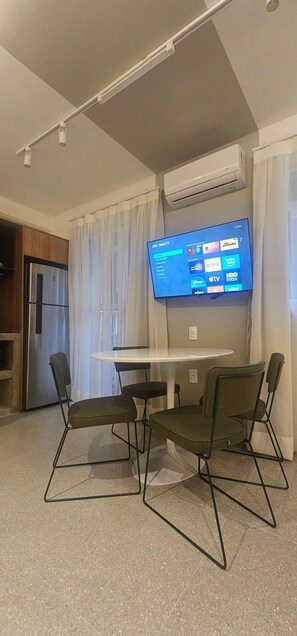 A room with a flat-screen TV mounted on the wall, a small table with chairs, a refrigerator, and an air conditioning unit. - Cond. Metro Casa Centro 101 (São Paulo)