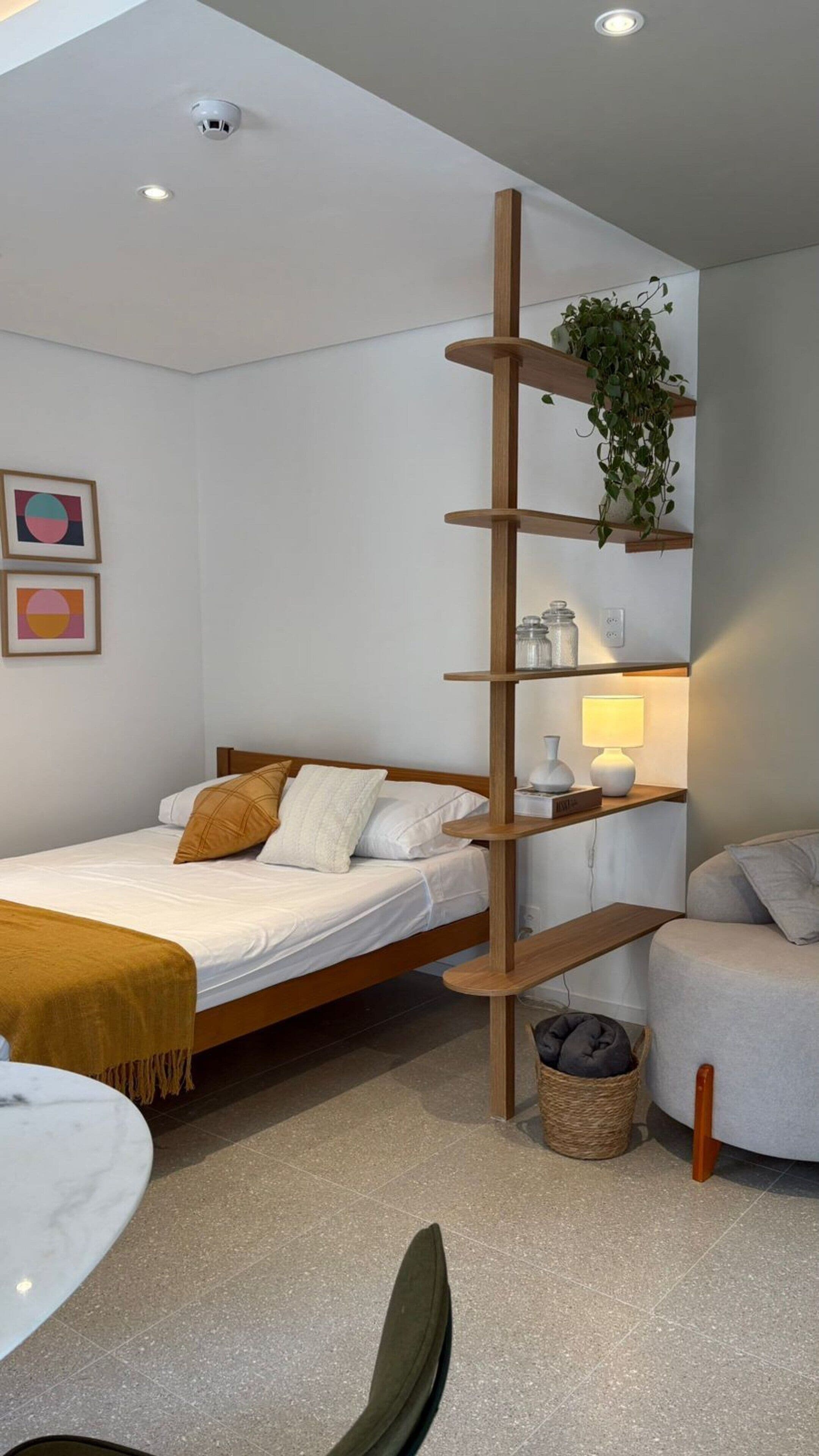 Een moderne slaapkamer met een bed, een nachtkastje met een lamp, een houten plank met een plant en een klein mandje.