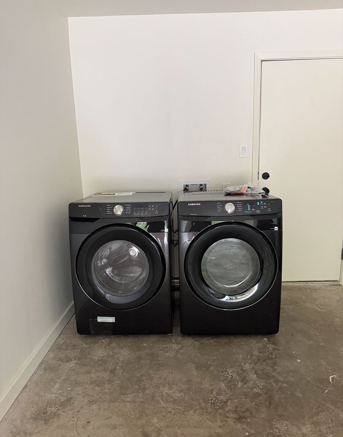 Deux lave-linge Samsung noirs à chargement frontal sont placés dans une pièce dont le mur est d’un blanc uni et où l’on aperçoit une porte fermée au fond.