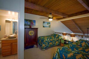 Ein Schlafzimmer mit Holzboden, Deckenventilator, zwei Betten mit tropischen Betttextilien, einem Holzschrank und einem Bild einer Strandlandschaft.