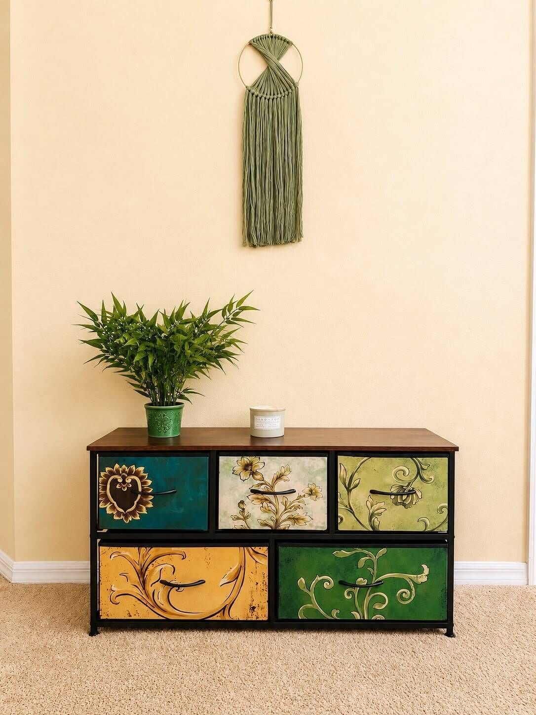 Un mueble de madera con diseños florales y abstractos, un colgador de macramé verde y una planta en maceta encima.