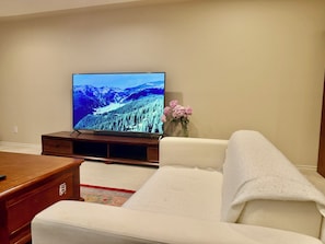 A living room with a flat-screen TV on a wooden stand, a white sofa, a small wooden coffee table, and a vase with pink flowers on a beige wall.