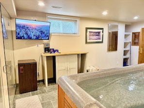 A bathroom with a shower, a toilet, a sink, a mirror, a towel rack, a window with blinds, a television mounted on the wall, and a framed picture on the wall.