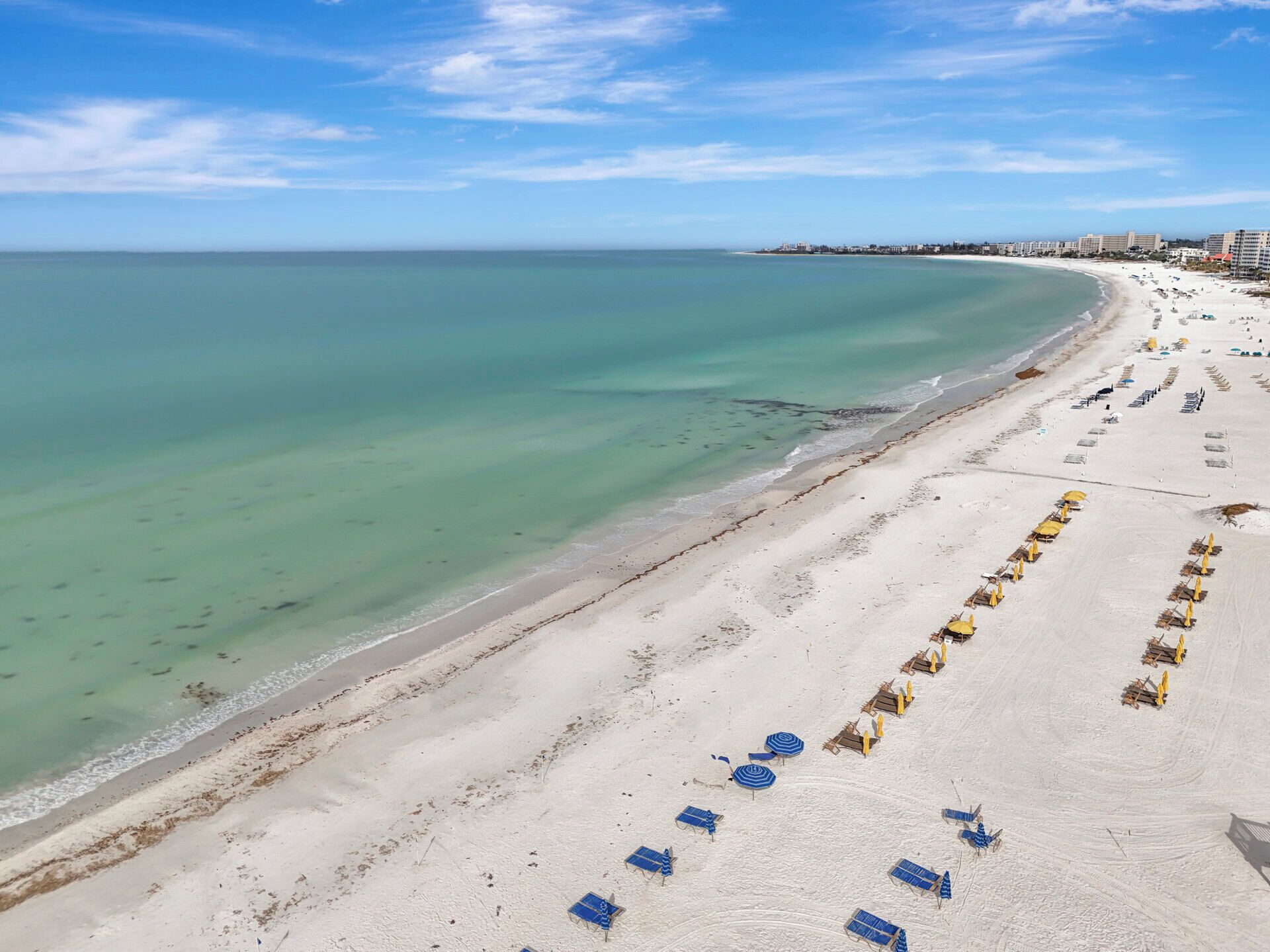 Une plage bordée de rangées de transats et de parasols, un ciel dégagé et une mer calme.