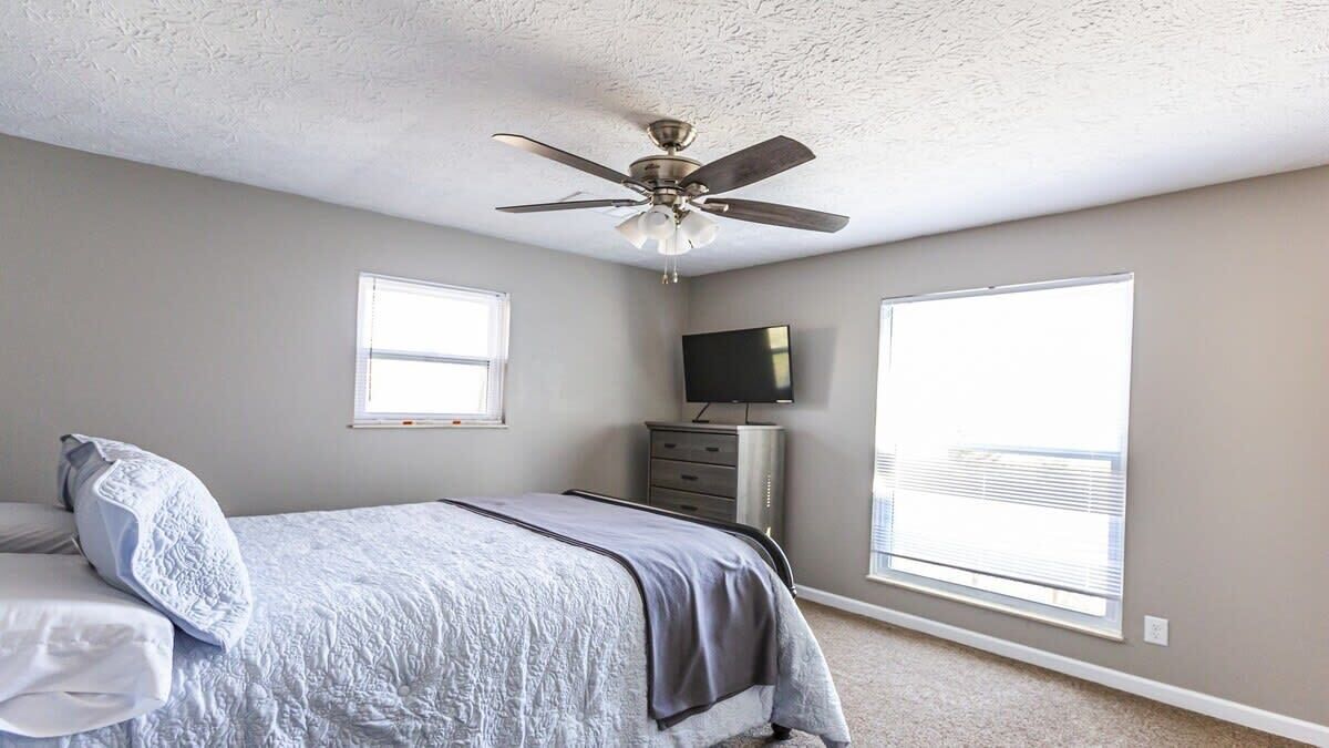 A bedroom with a bed, a ceiling fan, a dresser, a TV, and two windows.