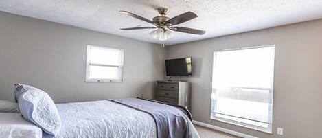Un dormitorio con cama, ventilador de techo, cómoda, televisor y dos ventanas.