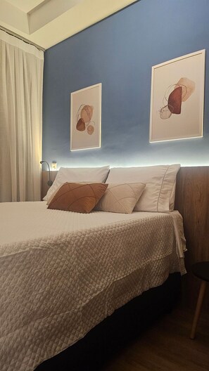A bedroom with a bed, two pillows, a headboard, a nightstand, a lamp, and two framed artworks on the wall. - Cond. Haus Mitre Reserva Vila Mariana  (São Paulo)