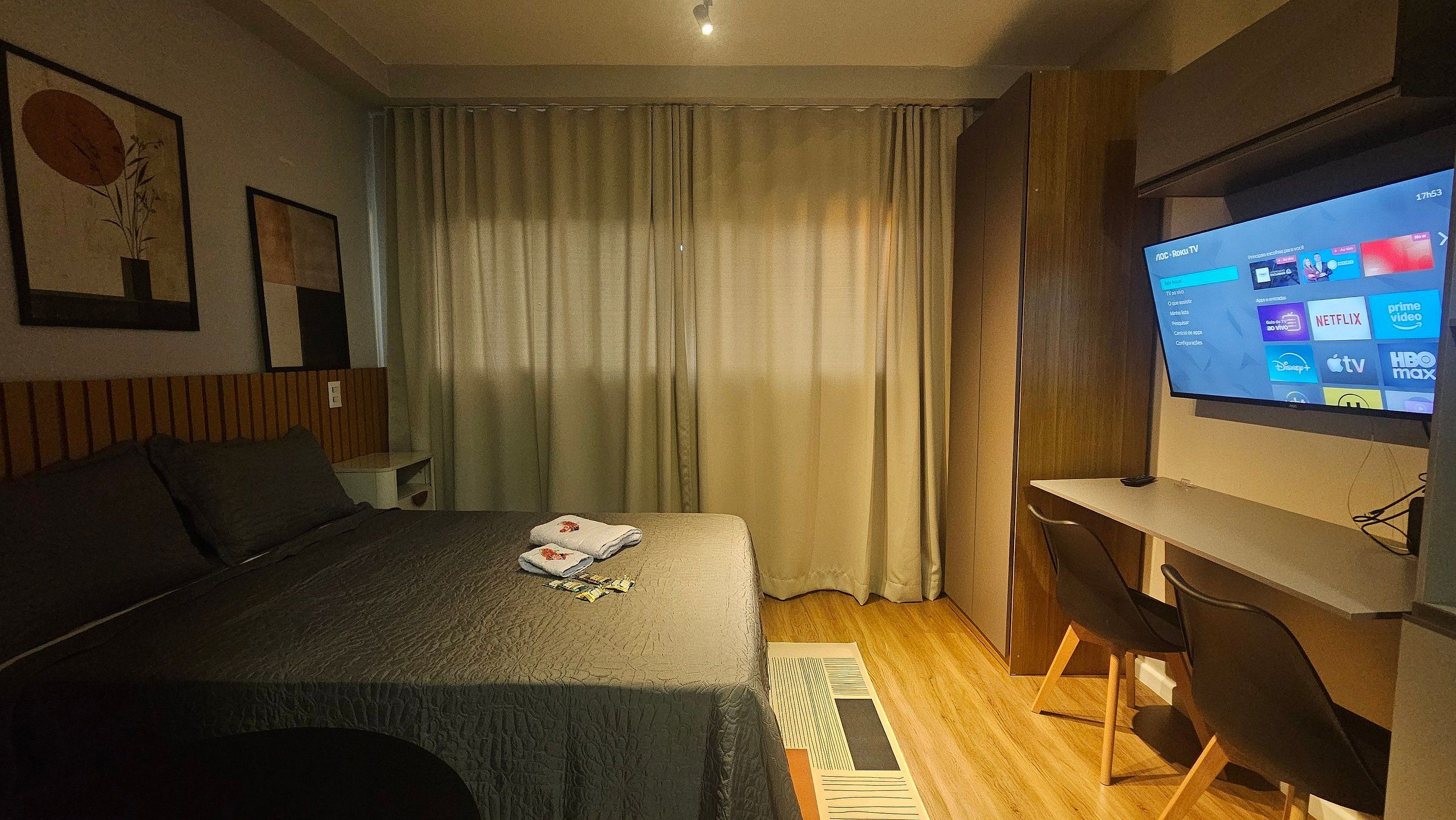 Habitación de hotel con cama, escritorio, televisor mostrando un menú y ventana con cortinas.