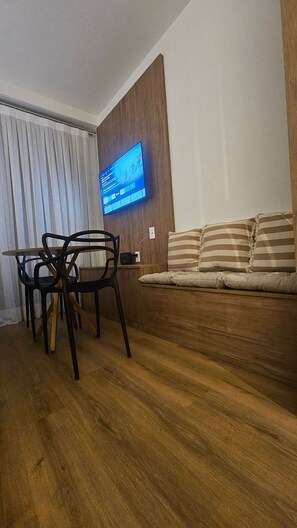 A living room with a wooden floor, a sofa with striped cushions, a television mounted on a wooden wall, and a dining table with chairs. - Cond. Haus Mitre Reserva Vila Mariana  (São Paulo)