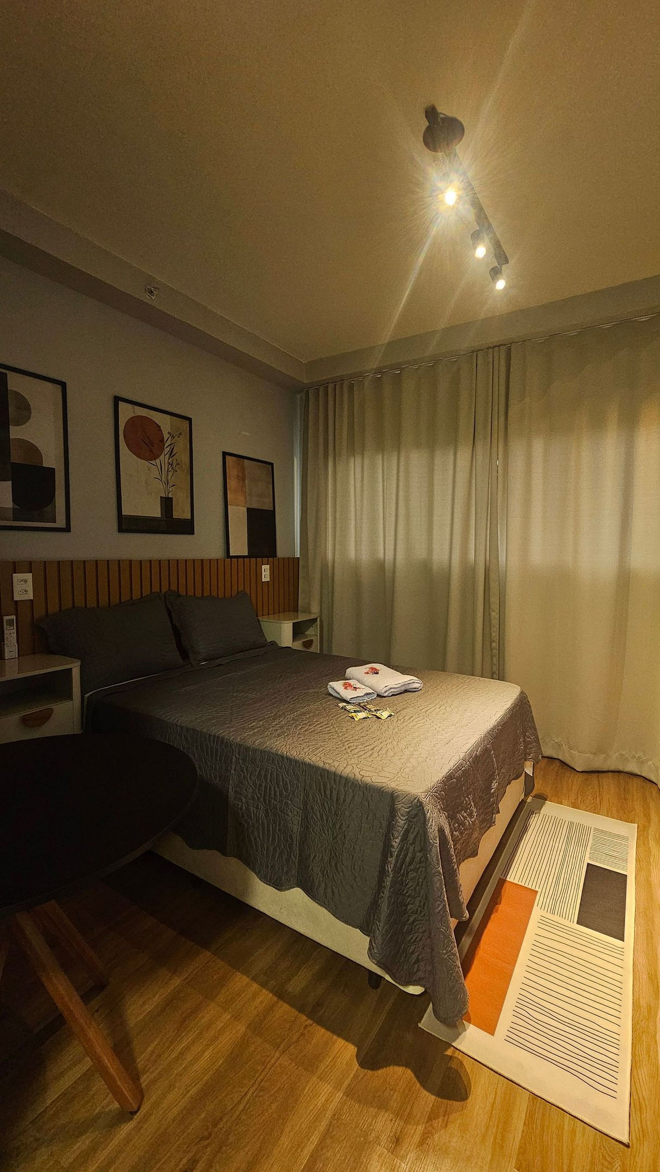 Quarto de hotel com cama, mesa de cabeceira, luminária, janela com cortinas e obra de arte na parede.