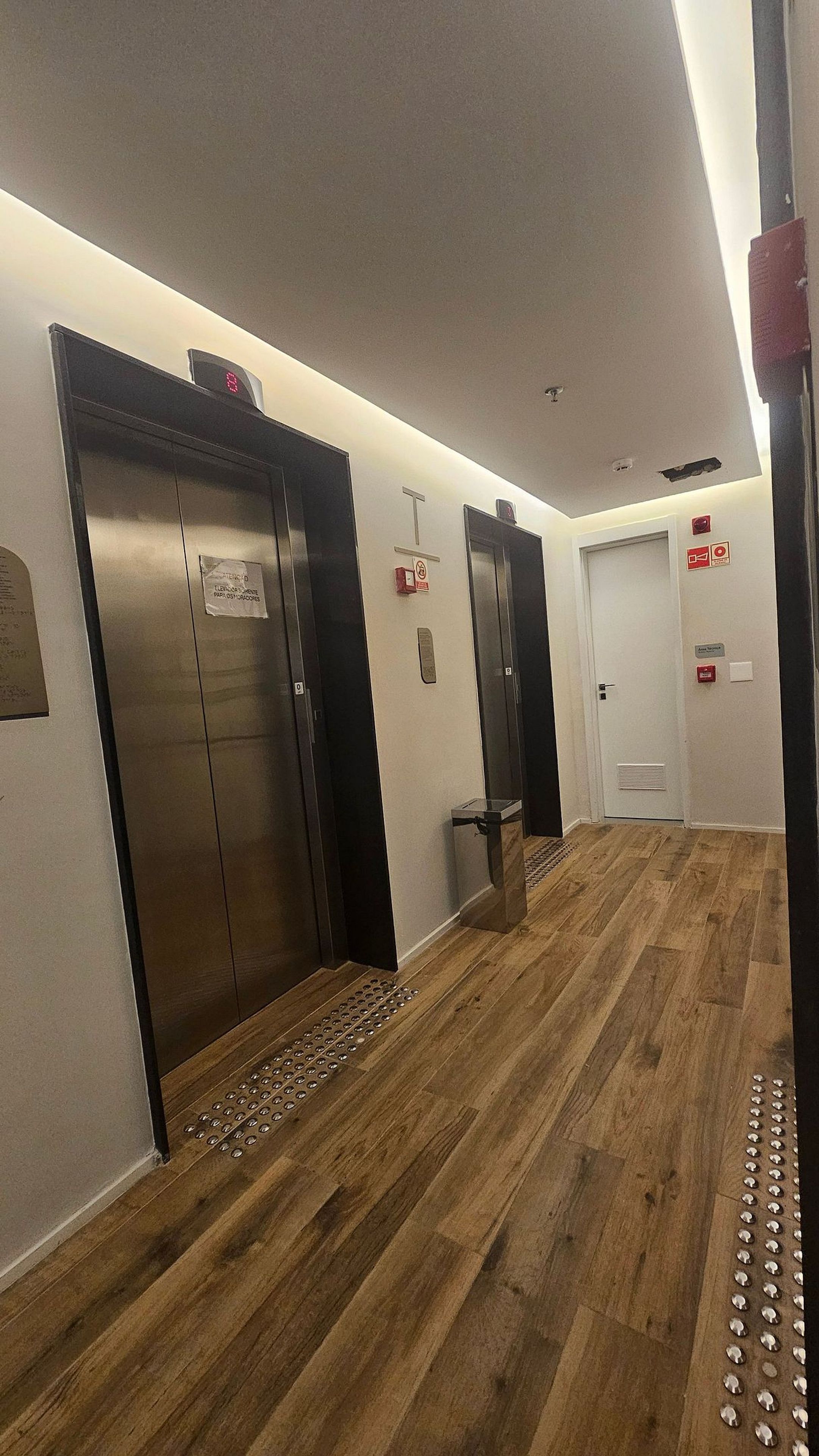 Un pasillo con suelo de madera, tres ascensores y una puerta con detector de incendios.