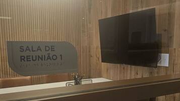 Uma sala de reuniões moderna com uma mesa comprida, duas cadeiras, uma TV de tela plana e um letreiro indicando 'Sala de Reunião 1'.