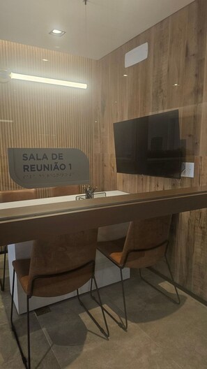 Uma sala de reuniões moderna com uma mesa comprida, duas cadeiras, uma TV de tela plana e um letreiro indicando 'Sala de Reunião 1'.