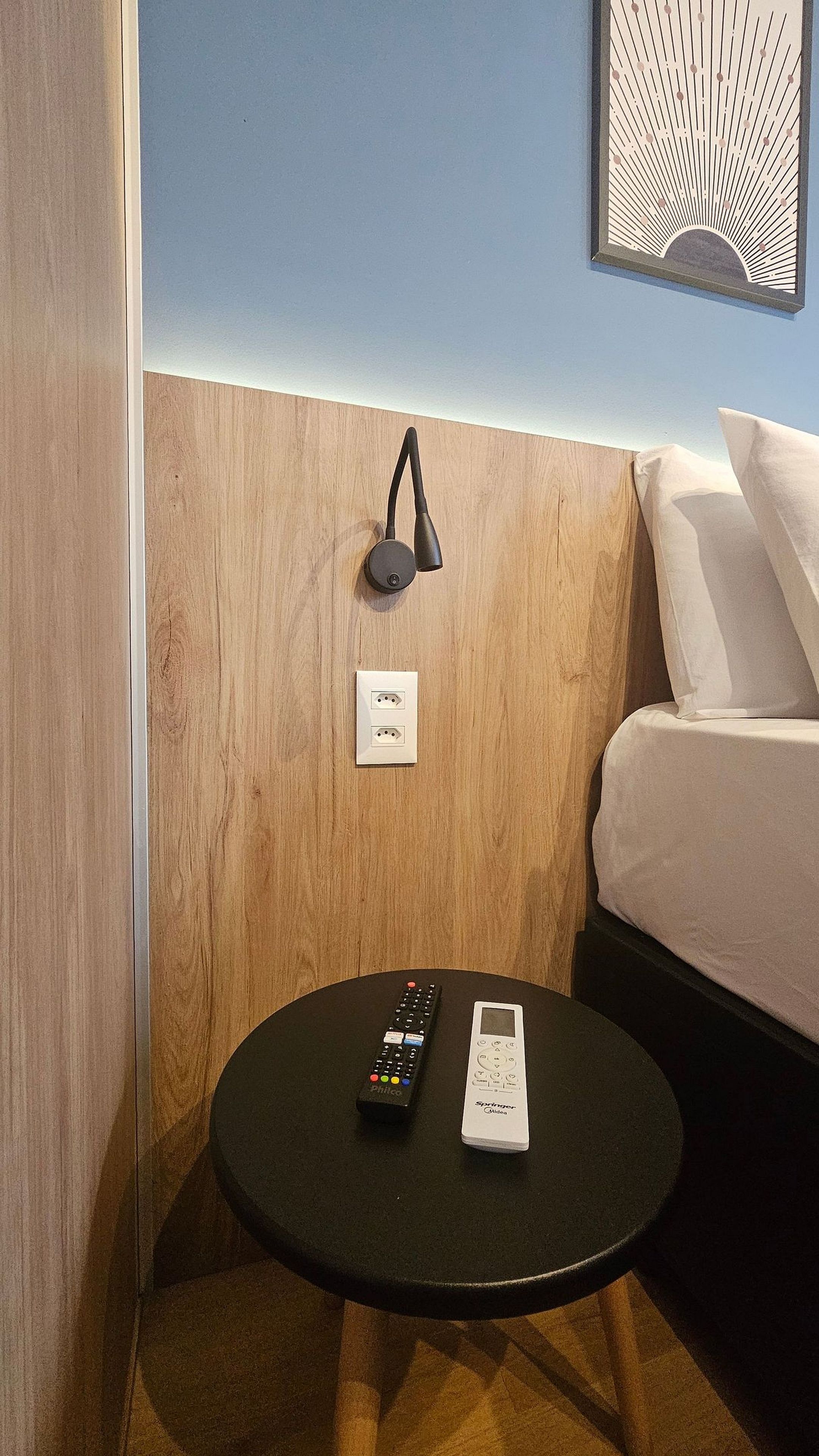 A hotel room with a bed, a bedside table, a remote control, a power outlet, and a wall-mounted lamp.