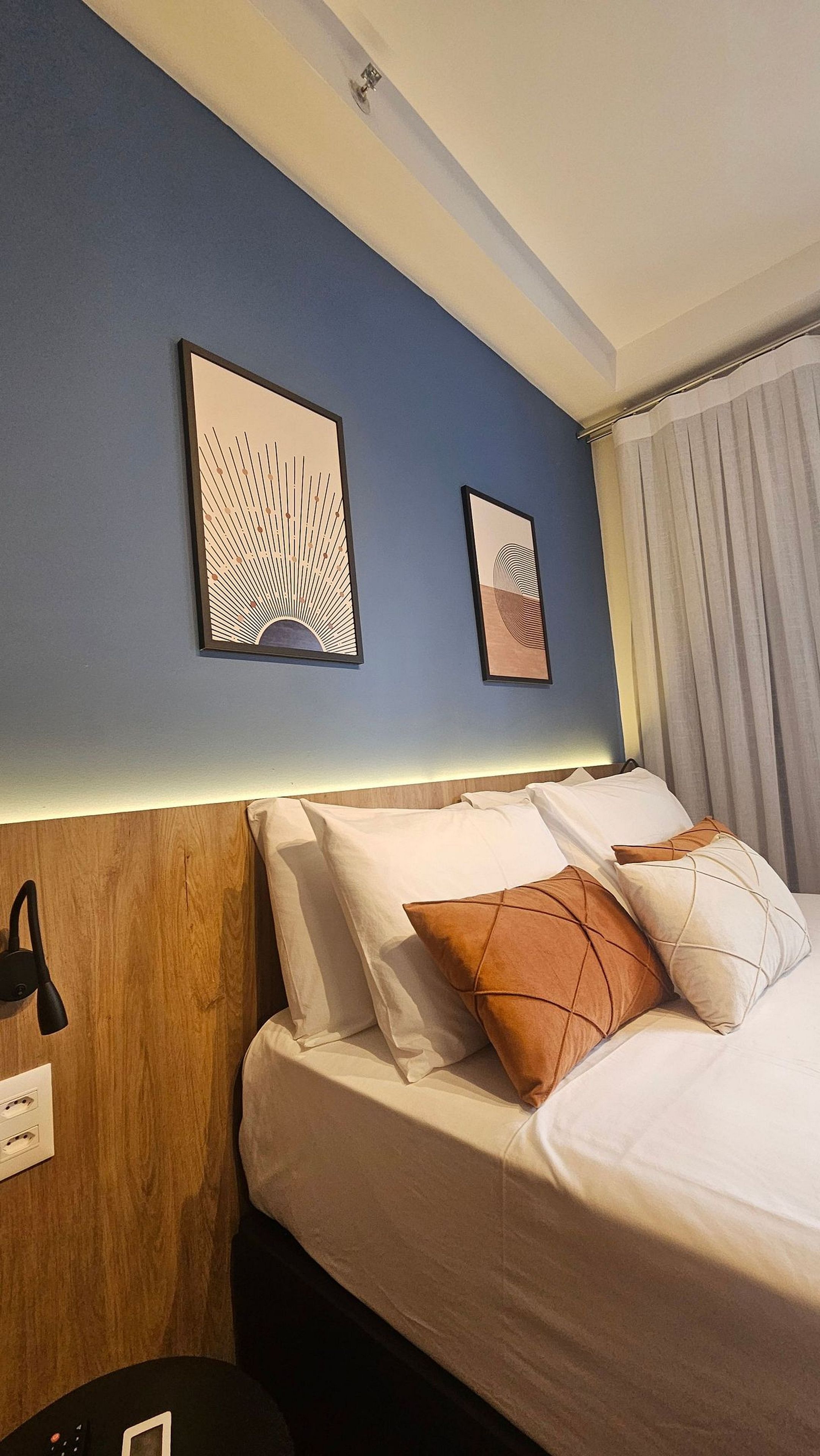 A hotel room with a bed, two wall-mounted artworks, a wooden headboard, and a nightstand with a lamp.