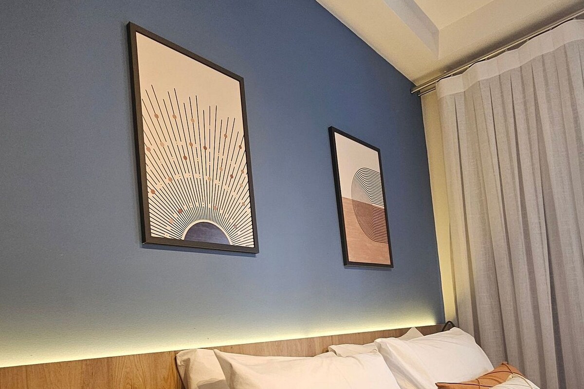 Quarto de hotel com uma cama, duas obras de arte penduradas na parede, cabeceira de madeira e mesa de cabeceira com uma luminária.