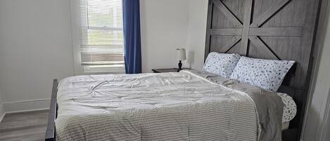 Un dormitorio con cama, mesita de noche, lámpara y ventana con cortinas.
