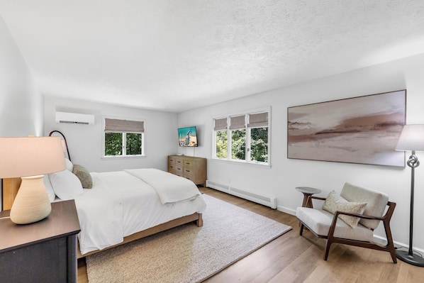 A bedroom with a bed, bedside table, lamp, dresser, and a large window with a view of trees.