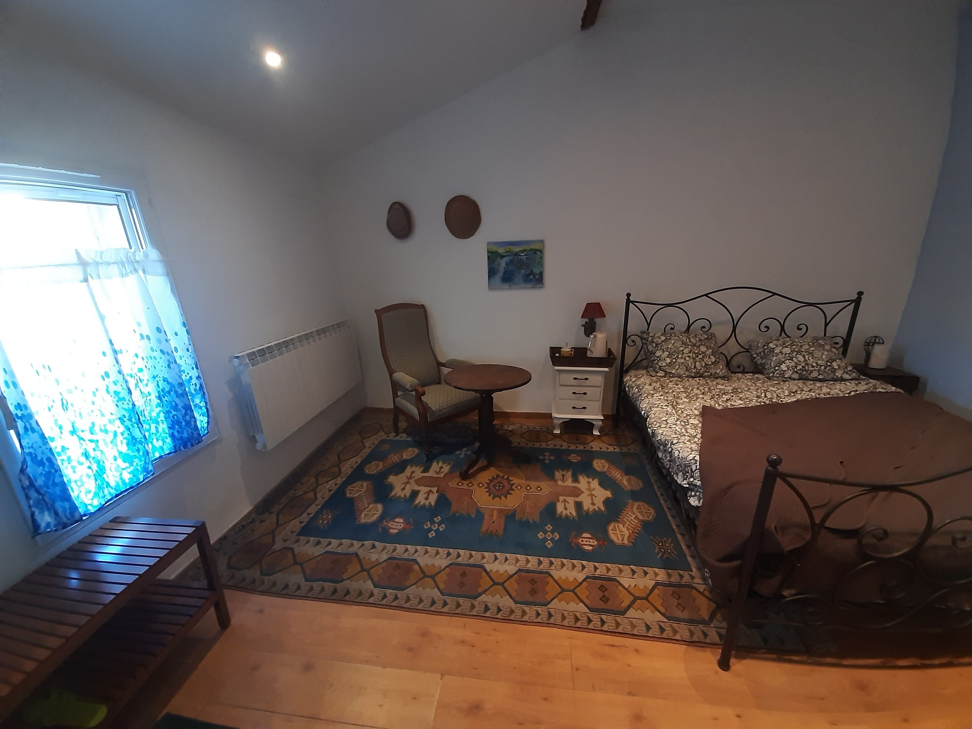 A bedroom with a bed, a chair, a small table, a radiator, and a window with curtains.