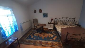 A bedroom with a bed, a chair, a small table, a radiator, and a window with curtains.