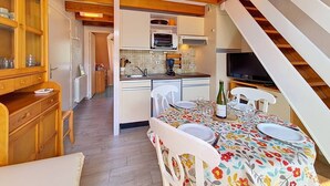 Una cocina compacta con vigas de madera, un área de comedor con mantel floral y un televisor sobre un pedestal.