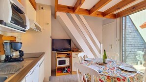 Cocina compacta con cafetera, microondas y televisor sobre una estantería de madera. Se ve un área de comedor con una mesa dispuesta para dos personas y mantel con motivos florales.