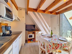 Eine kleine Küche mit Kaffeemaschine, Mikrowelle und Fernseher auf einem Holzboden. Ein Essbereich mit Tisch für zwei Personen und einer blumigen Tischdecke ist zu sehen.