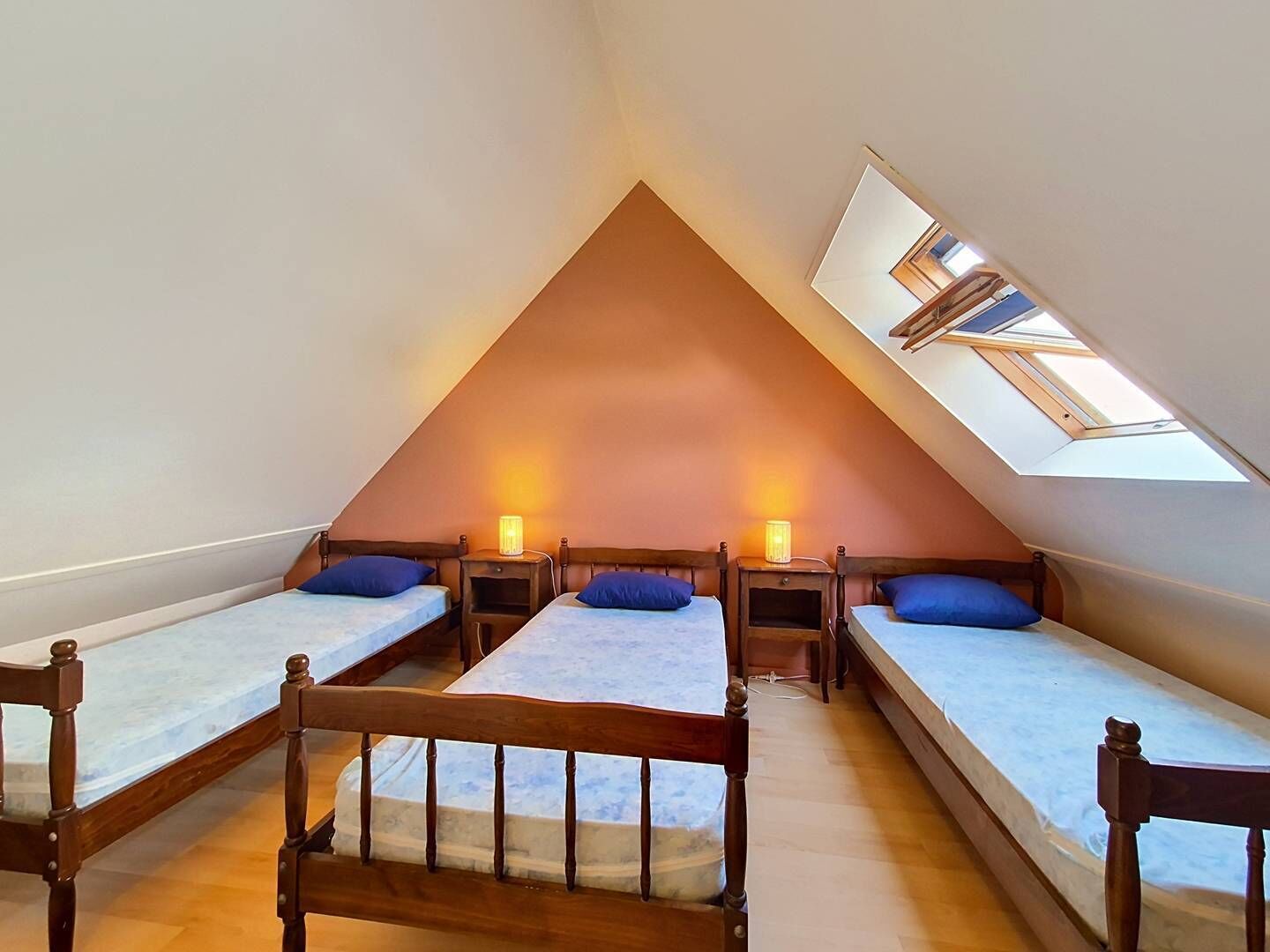 Een zolderkamer in driehoekige vorm met drie bedden, elk met een houten hoofdeinde, en een dakraam.