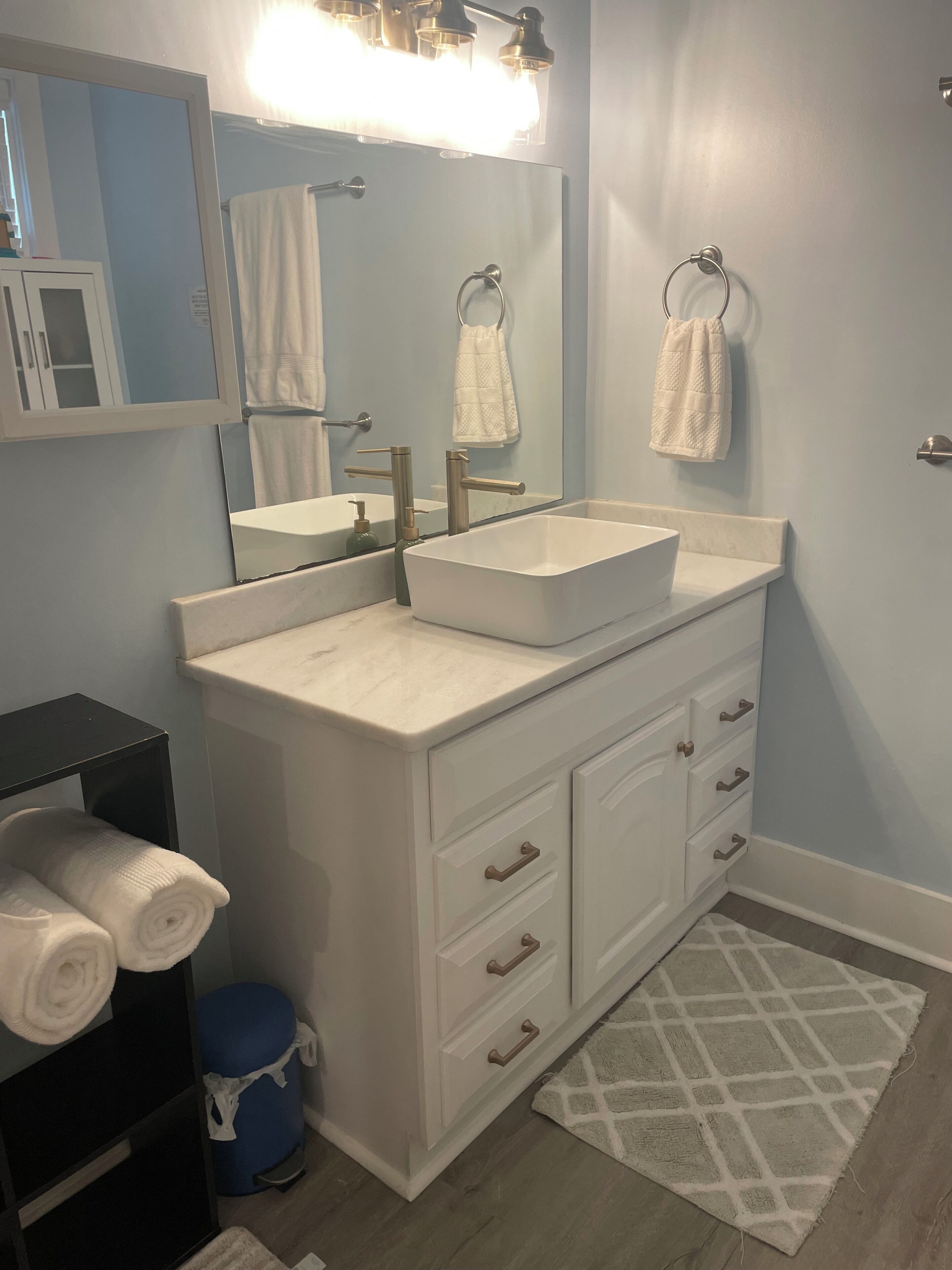 A bathroom with a double sink vanity, a mirror, towel racks, and a blue trash bin.