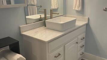 A bathroom with a double sink vanity, a mirror, towel racks, and a blue trash bin.