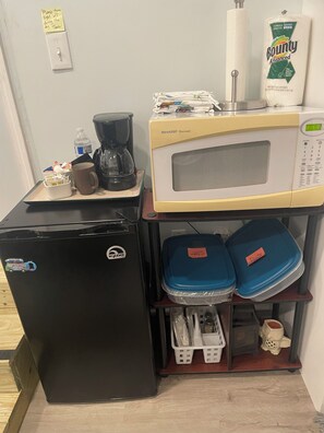 A compact kitchenette with a refrigerator, microwave, coffee maker, and storage containers.