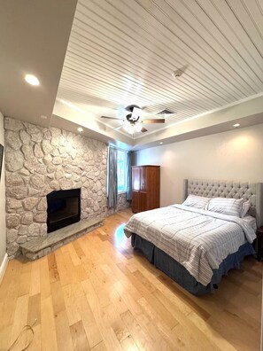 Une chambre avec une cheminée en pierre, un lit doté d’une tête de lit capitonnée et un ventilateur de plafond.