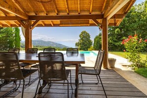 An outdoor wooden pergola with a table and chairs, overlooking a swimming pool and a mountainous landscape. - La Ferme de la Galoppaz (THOIRY)