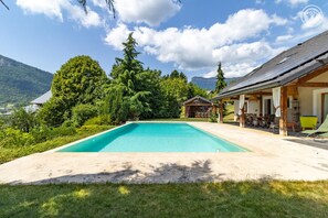 A swimming pool with a wooden deck, surrounded by lush greenery and a mountain view. - La Ferme de la Galoppaz (THOIRY)
