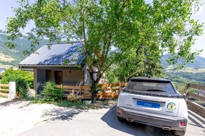 Ein weißer SUV vor einem rustikalen Gebäude mit einem Metall-Dach, umgeben von Grünflächen und Hügeln im Hintergrund.
