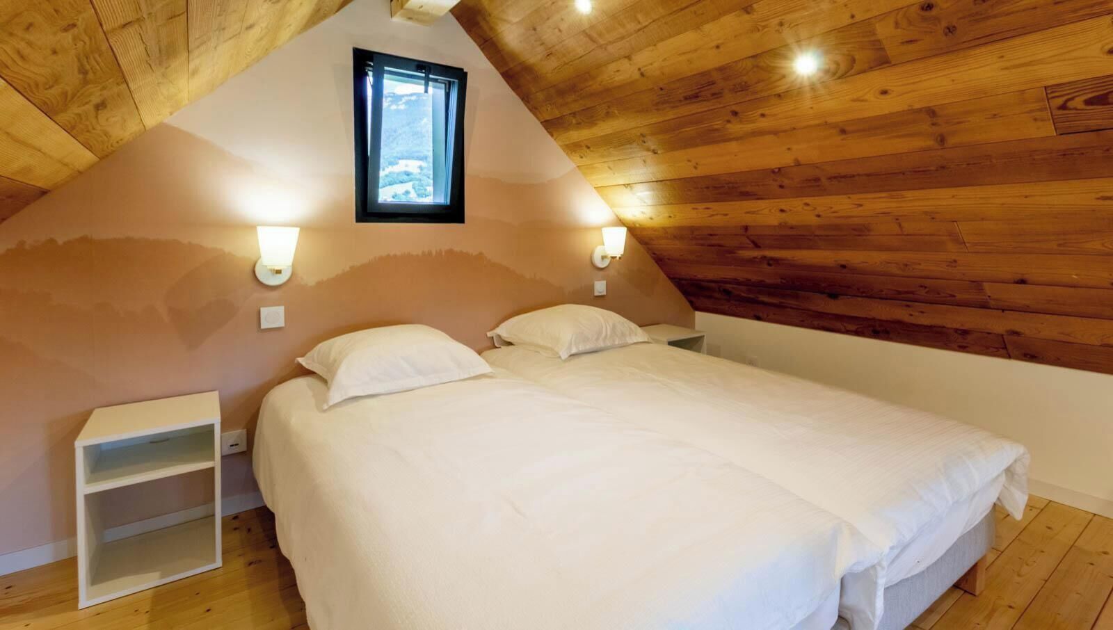 Une chambre avec un plafond en pente, un lit avec une literie blanche, deux tables de chevet et une fenêtre donnant sur un beau paysage.