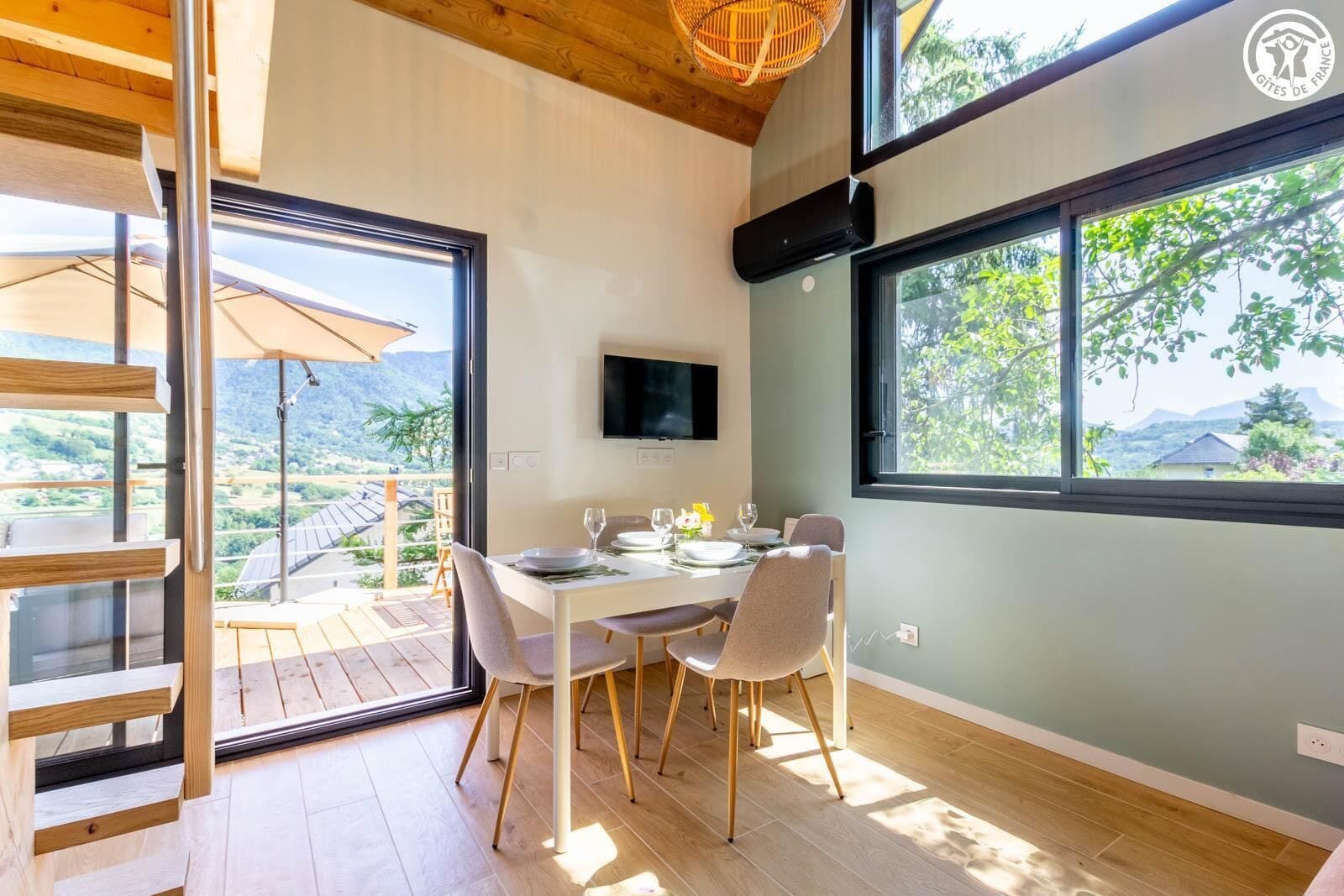 Comedor con una mesa dispuesta para cuatro, un televisor montado en la pared y un amplio ventanal con vistas al exterior.