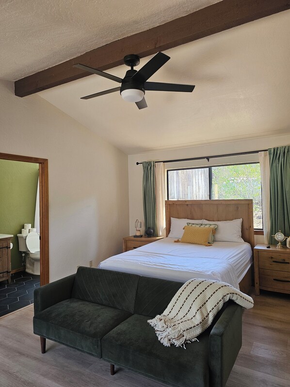 Une chambre à coucher avec un lit, une table de chevet, une commode, un ventilateur de plafond, une fenêtre avec des rideaux et un canapé vert.