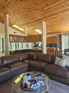 Ein modernes Wohnzimmer mit einem braunen Leder-Modulsofa, einem Glastisch mit Dekoration und einer Küche im Hintergrund.