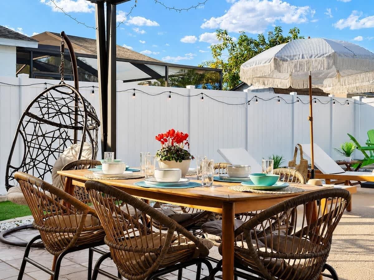 An outdoor dining area with wicker chairs, a wooden table, and a white fence in the background.
