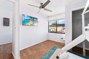 A room with a ceiling fan, a painting of a beach, a window with a view, and a white bed with a blue rug. - Heated Pool House Near Beach w/Game Room & Theater (Pinellas Park)