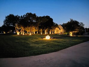 Uma área residencial bem iluminada ao entardecer, com uma casa, árvores e gramado.