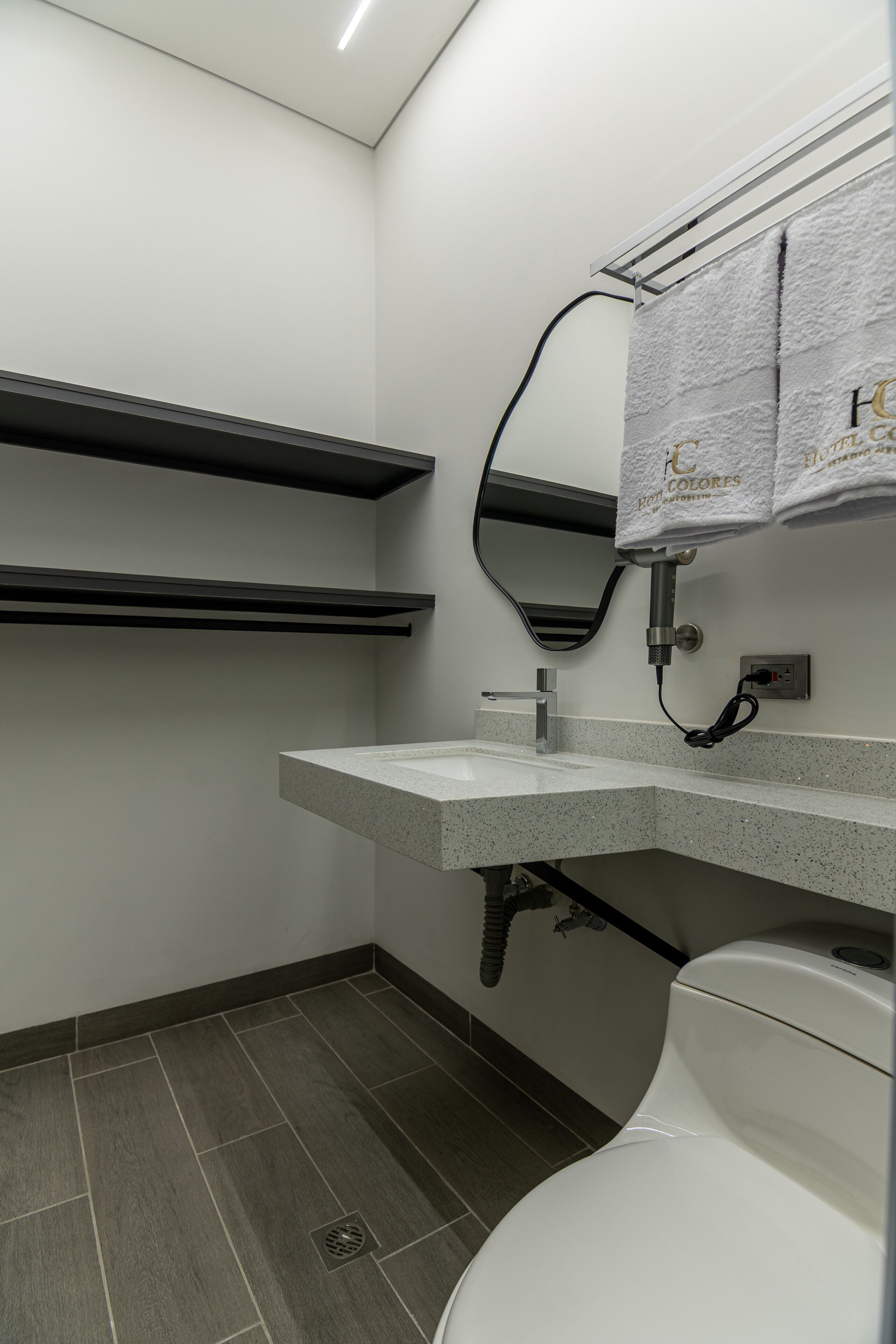 Un baño moderno con inodoro, lavamanos y espejo. Hay toallas colgadas en un perchero y un cabezal de ducha a la vista.