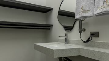 Un baño moderno con inodoro, lavamanos y espejo. Hay toallas colgadas en un perchero y un cabezal de ducha a la vista.