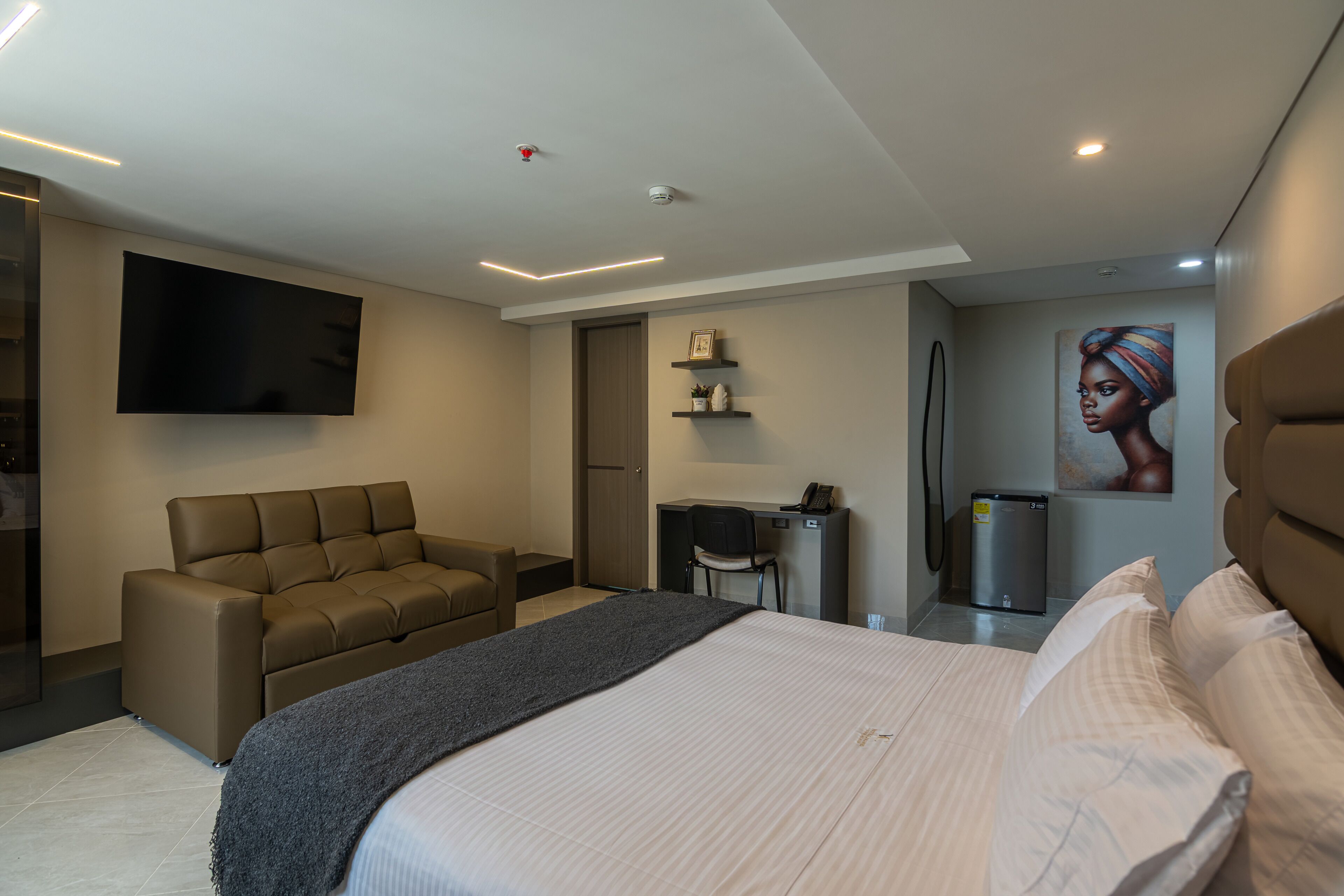 A modern hotel room with a large bed, a sofa, a desk with a chair, a flat-screen TV mounted on the wall, and a refrigerator.