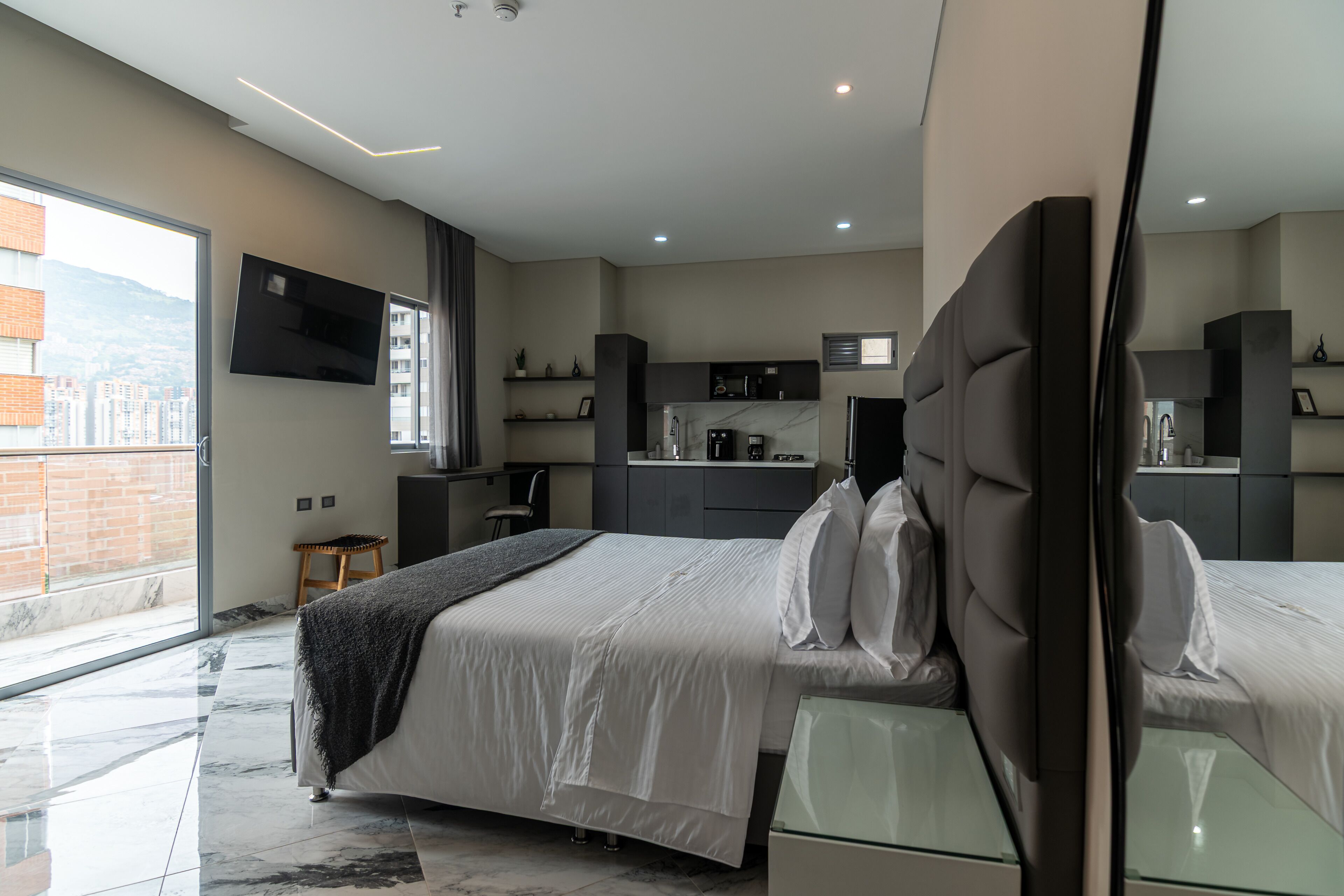A modern bedroom with a large bed, a small stool, a TV, and a kitchen area.