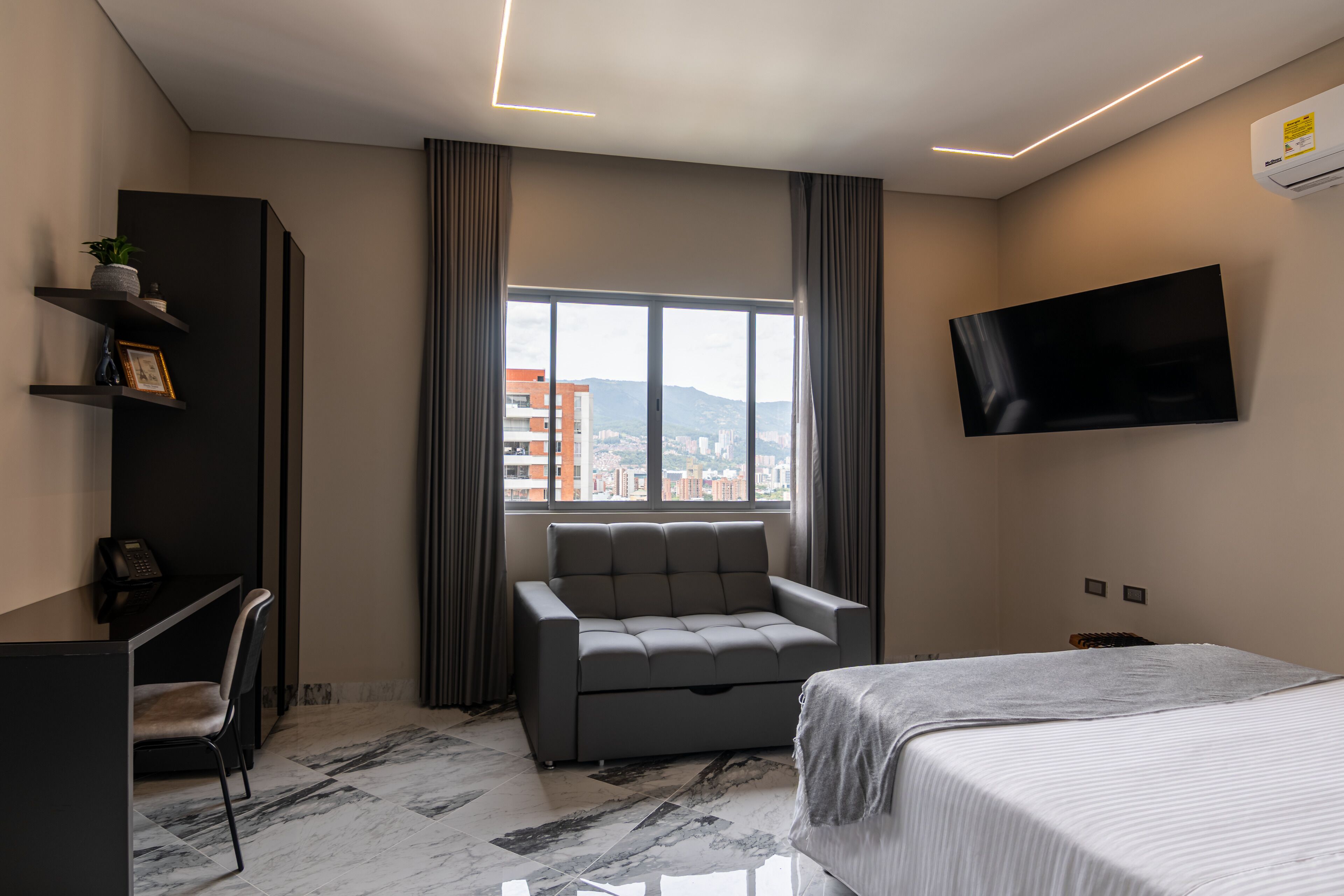 A modern hotel room with a bed, a grey sofa, a desk with a phone, a TV mounted on the wall, and a window with a city view.