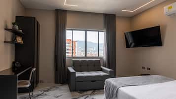 A modern hotel room with a bed, a grey sofa, a desk with a phone, a TV mounted on the wall, and a window with a city view.